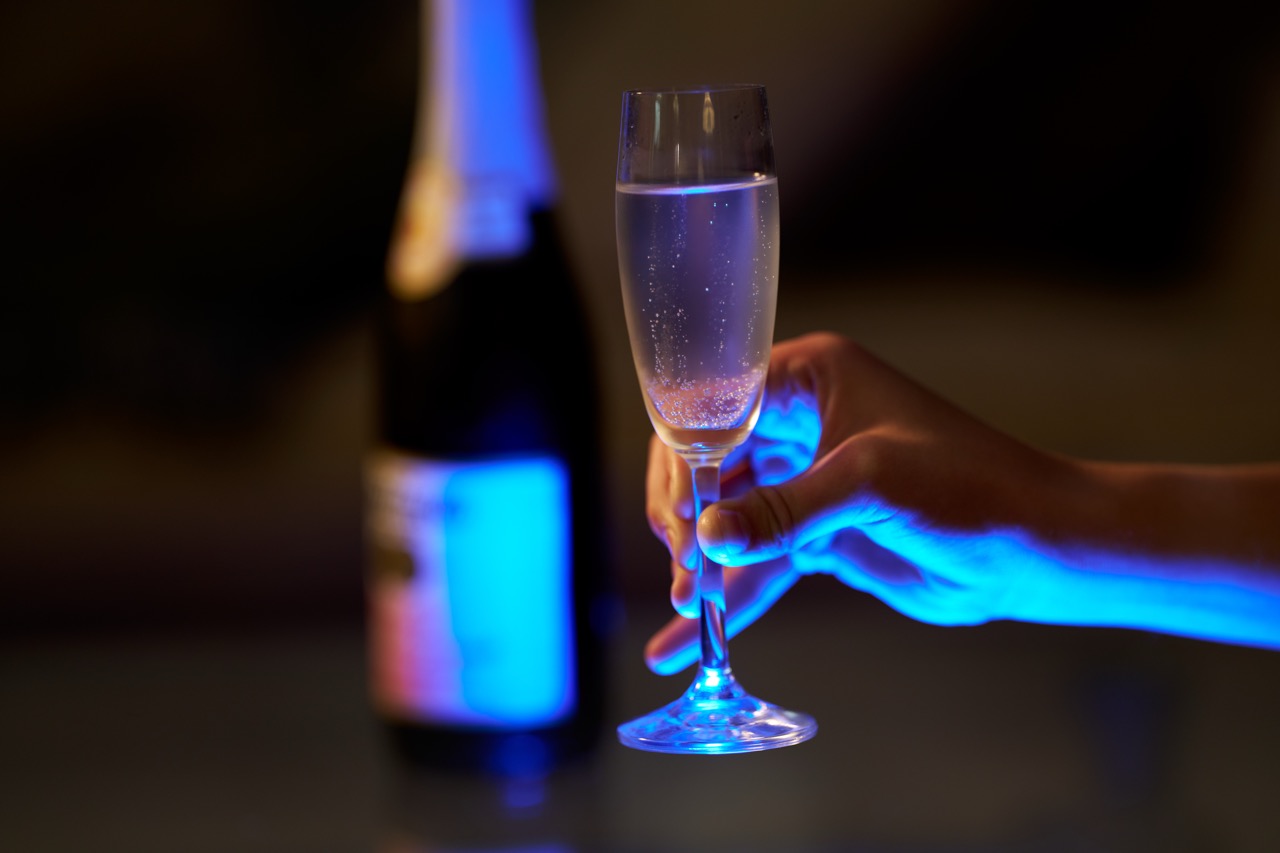 A hand holds a filled champaign glass while a bottle sits out of focus behind it.