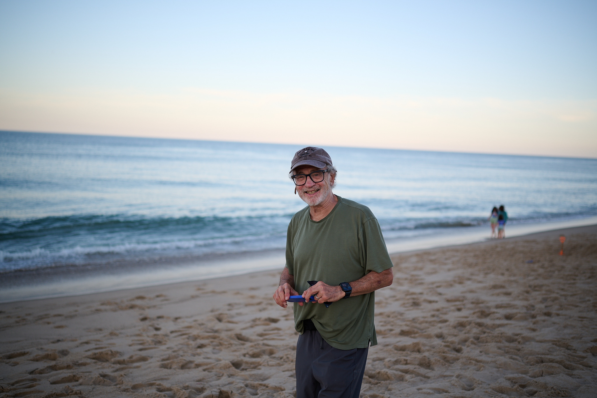 A portrait of a man on the beach. He's wearing a green shirt and puple cap, holding an iphone.